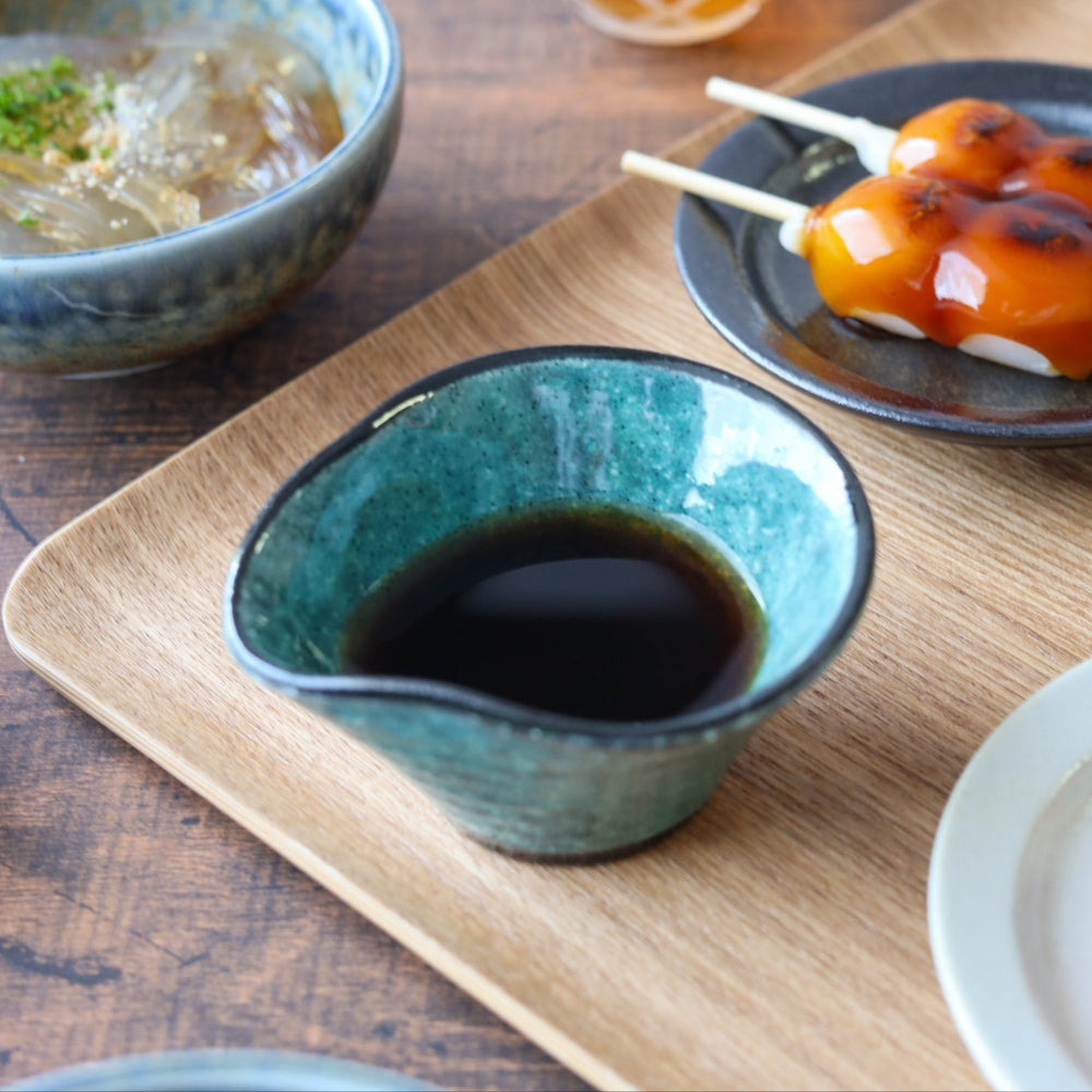 Wavy-edged blue-green condiment bowl filled with soy sauce, placed on a wooden tray alongside traditional Japanese sweets and chilled noodles.