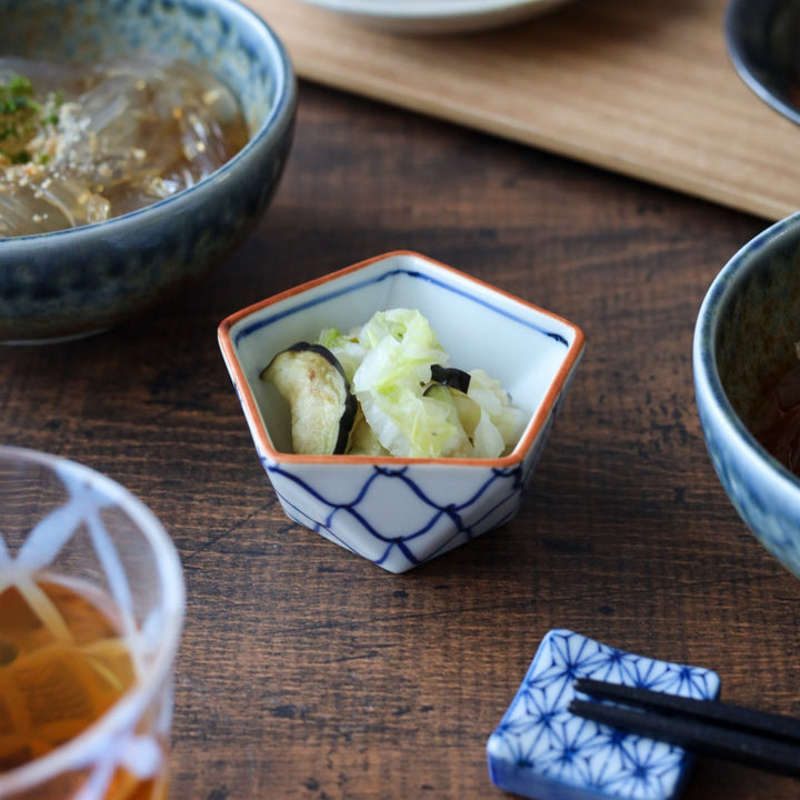 Pentagon-shaped porcelain condiment bowl with a blue mesh pattern and orange rim, beautifully styled with pickled vegetables on a wooden table.