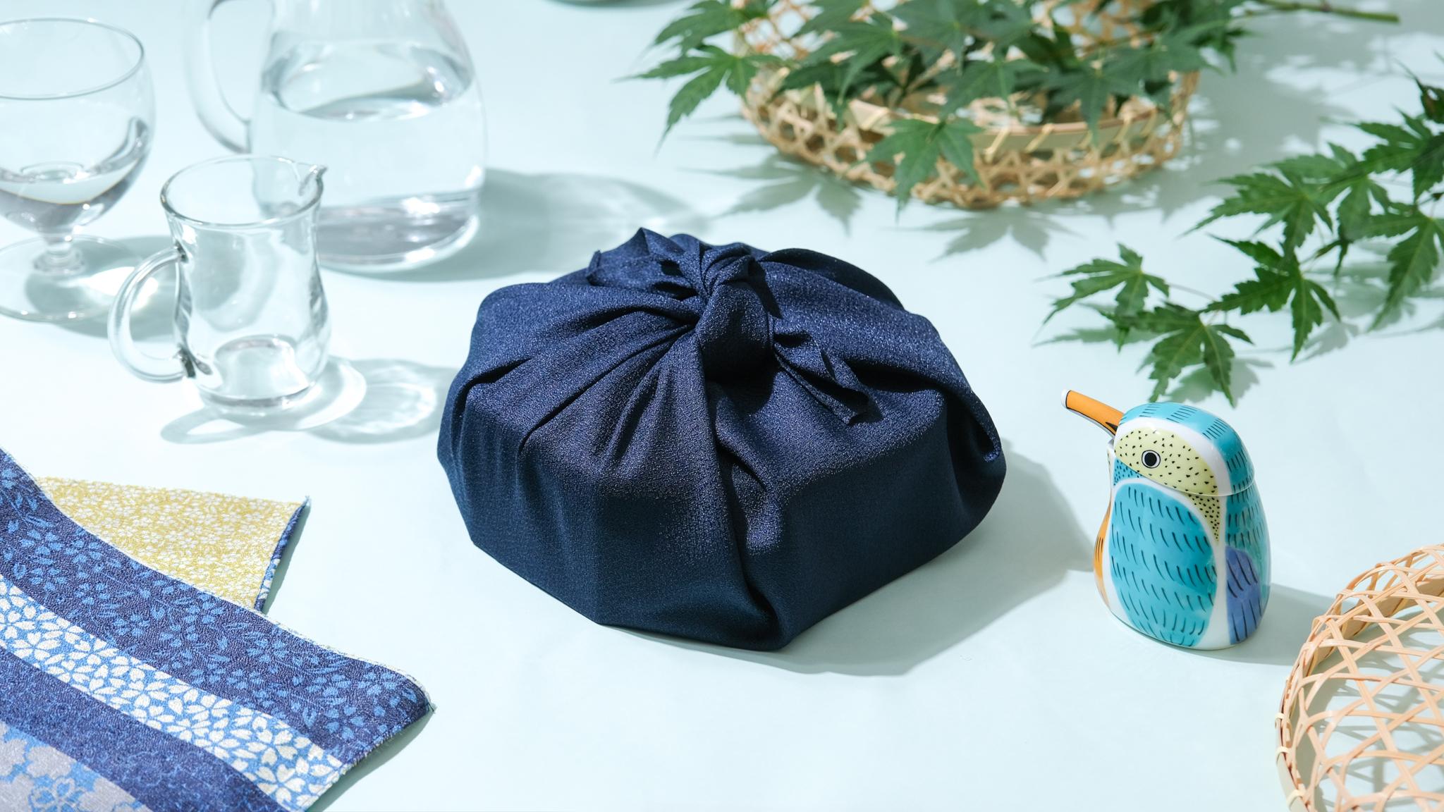 Products wrapped in a navy Japanese furoshiki cloth, tied in a knot on top, next to glass cups and folded patterned furoshiki.