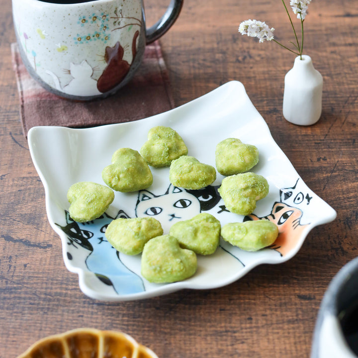 Wavy-edged white dessert plate featuring illustrated cats in black, white, and orange, playfully arranged beneath green heart-shaped treats.
