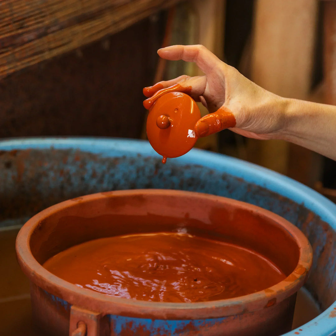 Artisan lifting a clay teapot lid from red slip to coat it evenly during traditional hand crafted pottery work