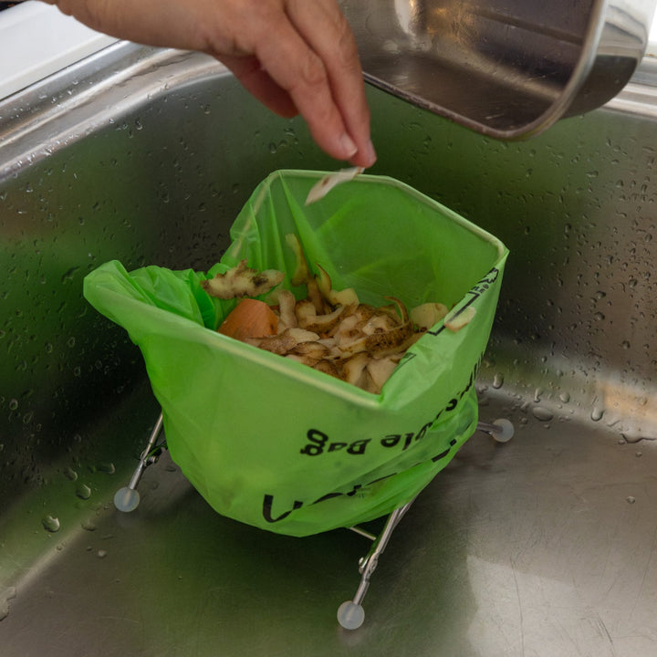 Nami throwing veggie peels into ecoframe stainless compost holder inside kitchen sink food waste bag stand