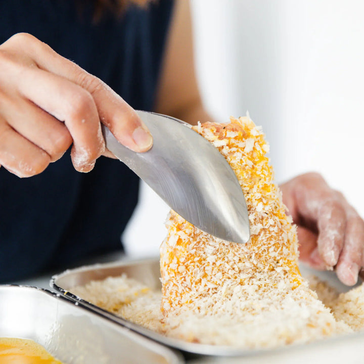 Close up of AUX Japanese Easy Tongs holding a pork cutlet as it is coated in breadcrumbs in a tray