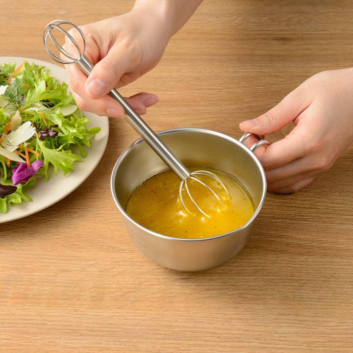 Person whisking salad dressing with a mini whisk inside a stainless steel egg beating bowl with handle