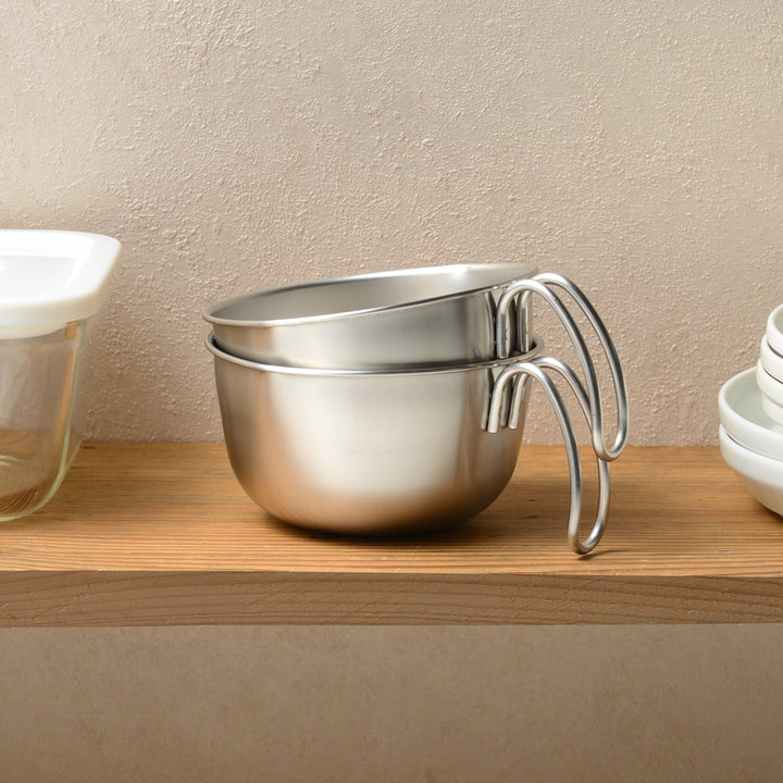 Two stainless steel egg beating bowls with handles neatly stacked for storage on a wooden shelf