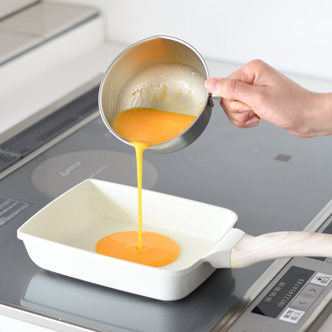 Person pouring beaten egg mixture from a stainless steel egg beating bowl with handle into a square tamagoyaki pan