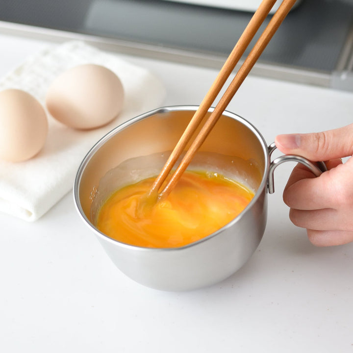 Person beating raw eggs with wooden chopsticks inside a stainless steel egg beating bowl with handle