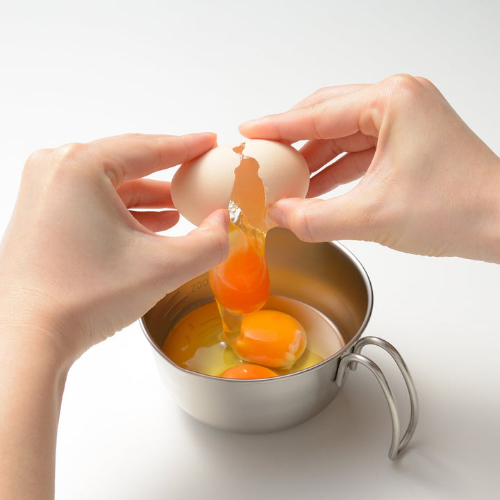 Person cracking a raw egg into a stainless steel egg beating bowl with handle containing other eggs