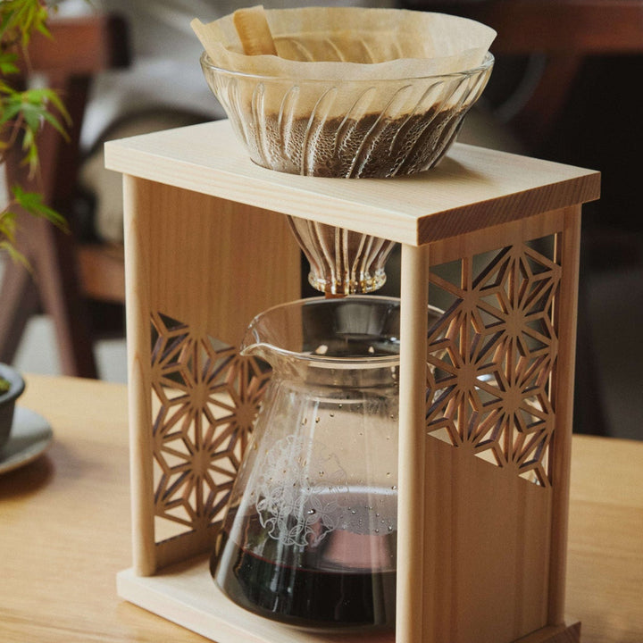 Freshly brewed coffee inside aya asanoha wood coffee dripper set sitting next to green bonsai tree on wooden table