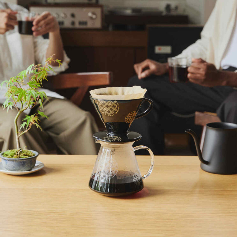 Displaying black hario ceramic coffee dripper on glass server with bonsai tree and friends drinking coffee in background