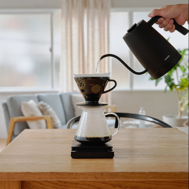 Pouring hot water from black kettle into hario ceramic coffee dripper and glass server on digital scale in living room