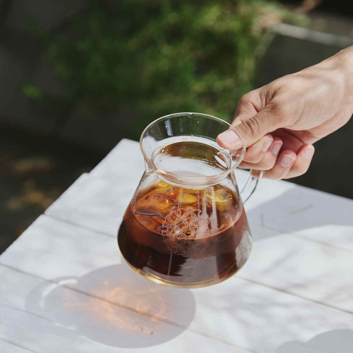 Holding hario patterned glass range server with iced coffee casting colorful shadow on white wooden table outdoors
