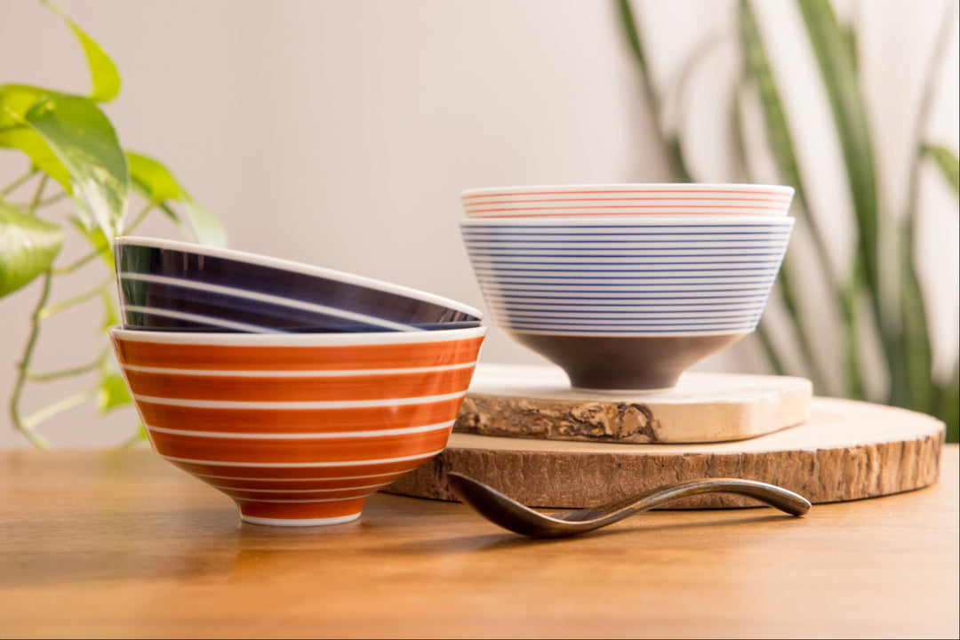 Colorful striped Japanese rice bowls in red, blue, and white arranged on a wooden table with plants in the background.