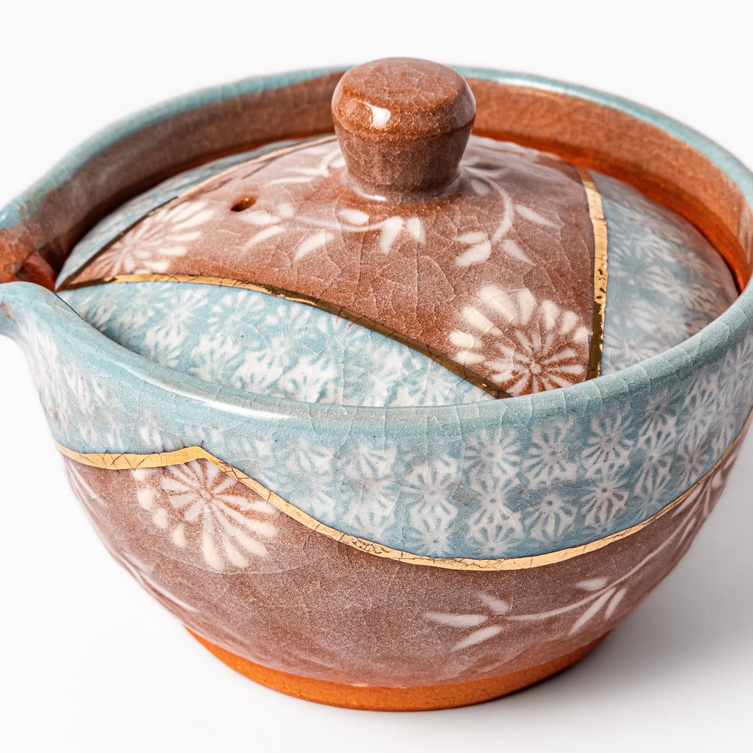 Ceramic teapot with lid featuring a turquoise crackle glaze and floral patterns in soft brown tones accented by a curved gold line.