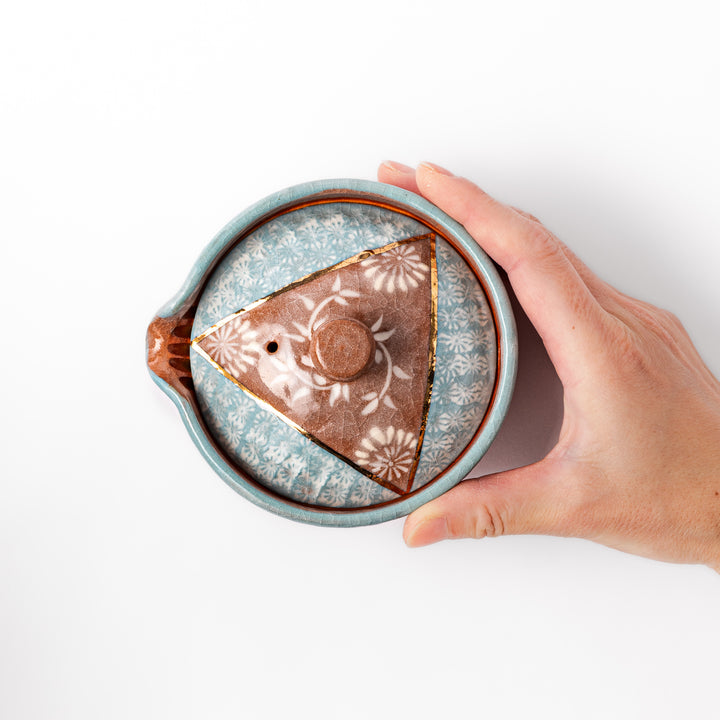 Ceramic teapot with lid featuring a turquoise crackle glaze and floral patterns in soft brown tones accented by a curved gold line.