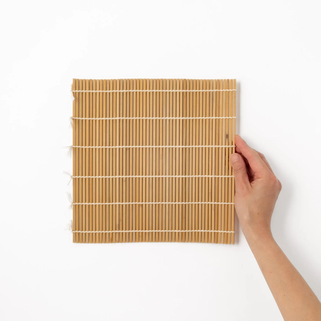 Traditional bamboo sushi rolling mat with uniform slats and white cotton string, partially rolled to show flexibility for making perfect sushi at home.