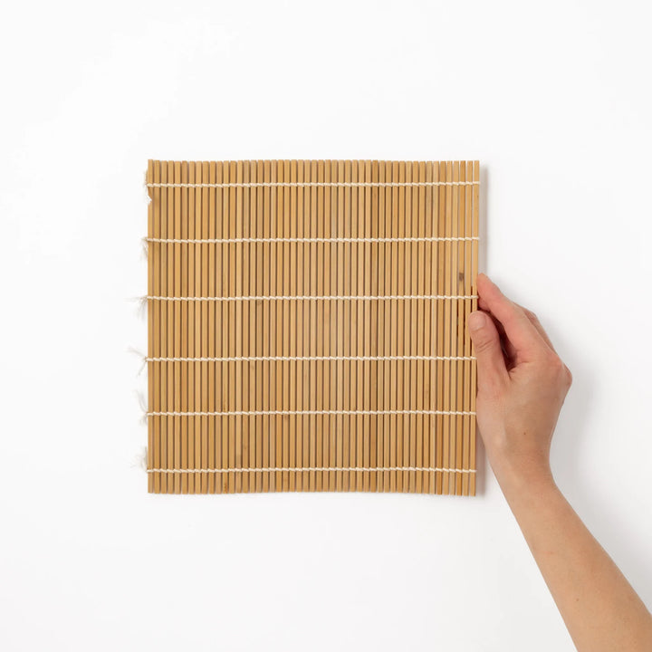 Traditional bamboo sushi rolling mat with uniform slats and white cotton string, partially rolled to show flexibility for making perfect sushi at home.