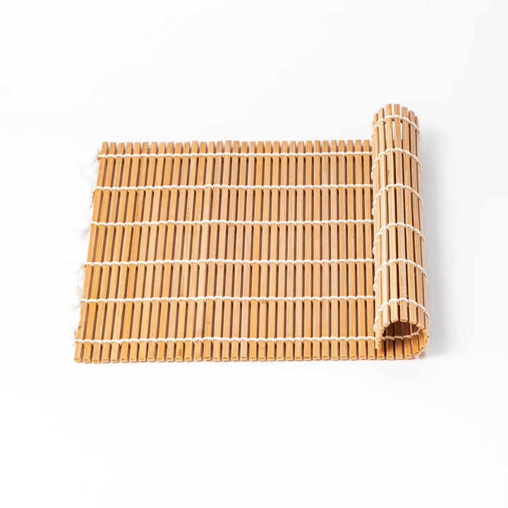 Traditional bamboo sushi rolling mat with uniform slats and white cotton string, partially rolled to show flexibility for making perfect sushi at home.