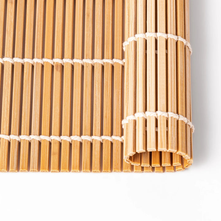 Traditional bamboo sushi rolling mat with uniform slats and white cotton string, partially rolled to show flexibility for making perfect sushi at home.