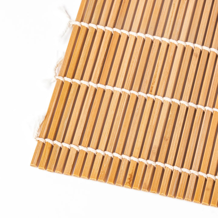 Traditional bamboo sushi rolling mat with uniform slats and white cotton string, partially rolled to show flexibility for making perfect sushi at home.