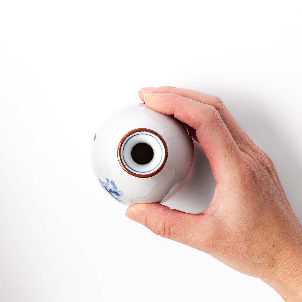 Porcelain sake flask with hand-painted blue mountain and village scenery, featuring a red rim and elegant traditional silhouette.