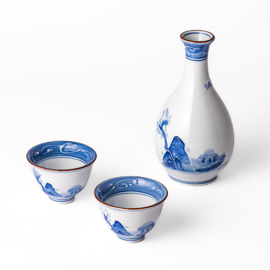 Three piece porcelain sake set with blue hand-painted mountain scenery, featuring a tall flask and two matching cups with red rims.