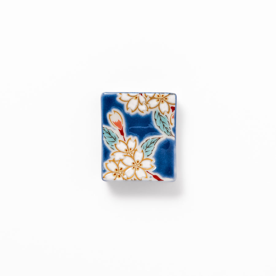 Rectangular blue Japanese ceramic chopstick rest with delicate white sakura blossoms and green leaves, ideal for traditional dining.