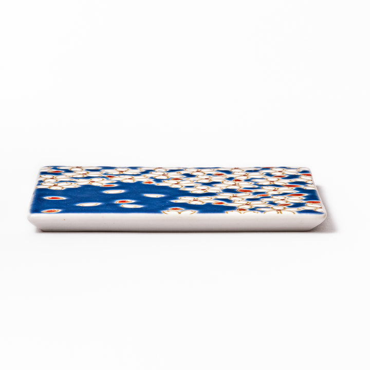 Square blue Japanese ceramic coaster with delicate white sakura blossoms and scattered petals, perfect for traditional dining settings.