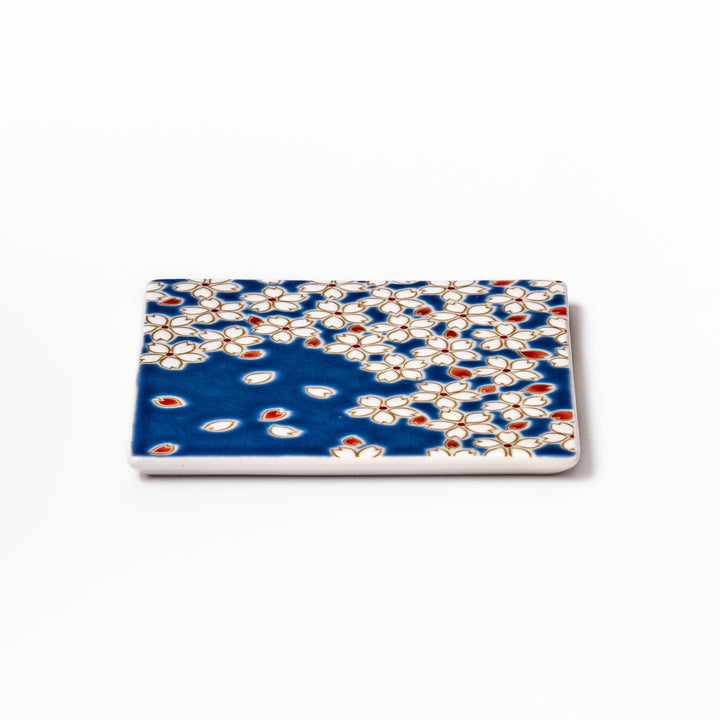 Square blue Japanese ceramic coaster with delicate white sakura blossoms and scattered petals, perfect for traditional dining settings.