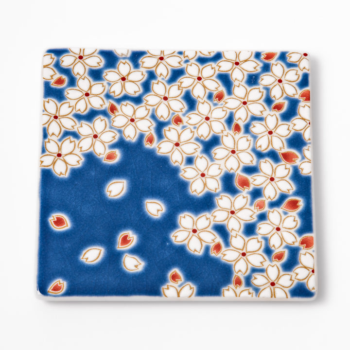 Square blue Japanese ceramic coaster with delicate white sakura blossoms and scattered petals, perfect for traditional dining settings.
