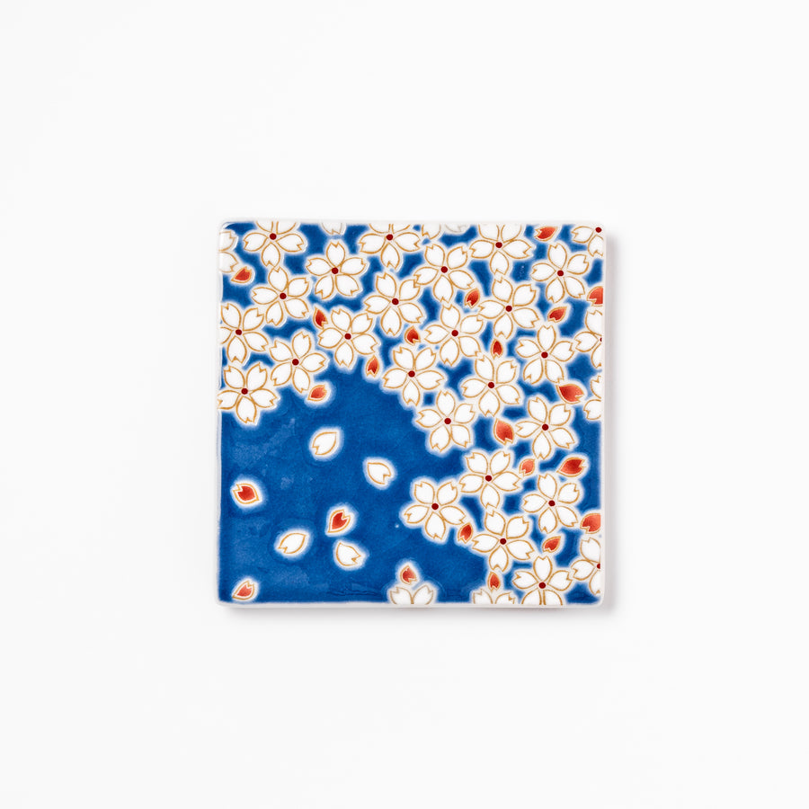 Square blue Japanese ceramic coaster with delicate white sakura blossoms and scattered petals, perfect for traditional dining settings.