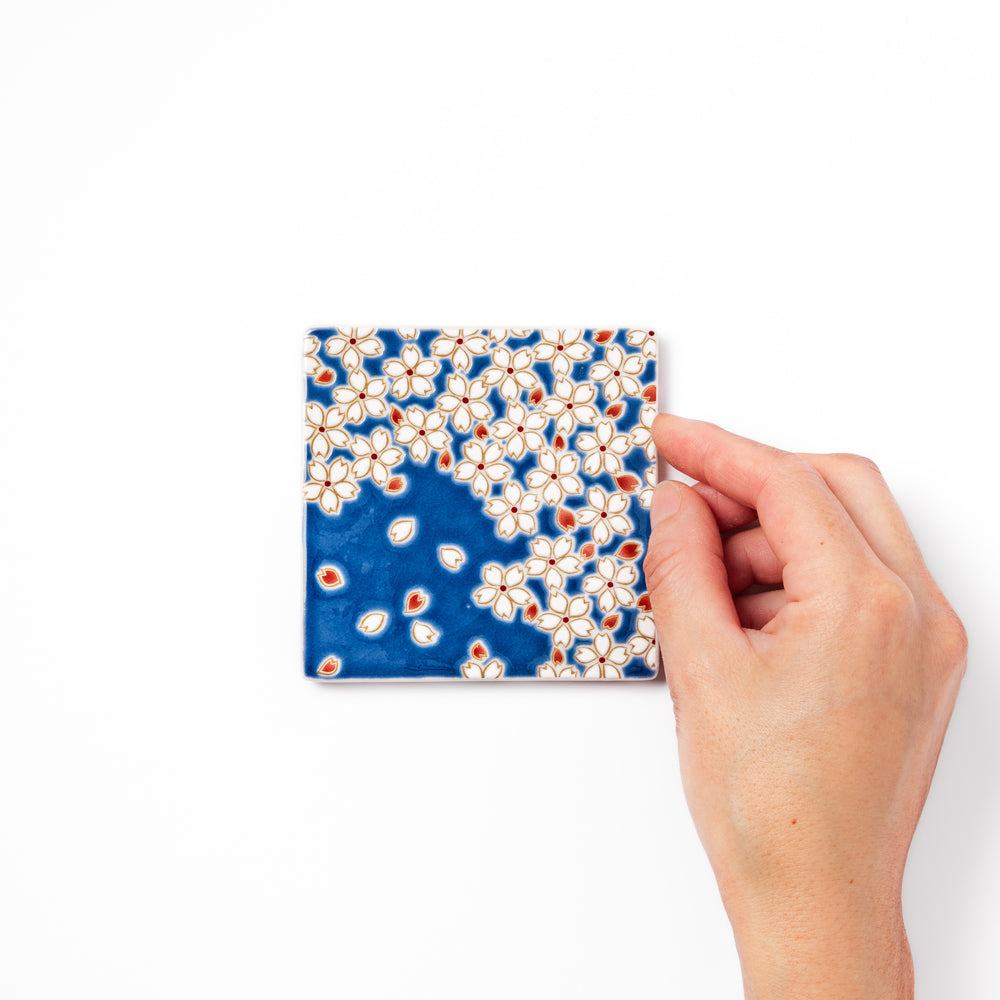 Square blue Japanese ceramic coaster with delicate white sakura blossoms and scattered petals, perfect for traditional dining settings.