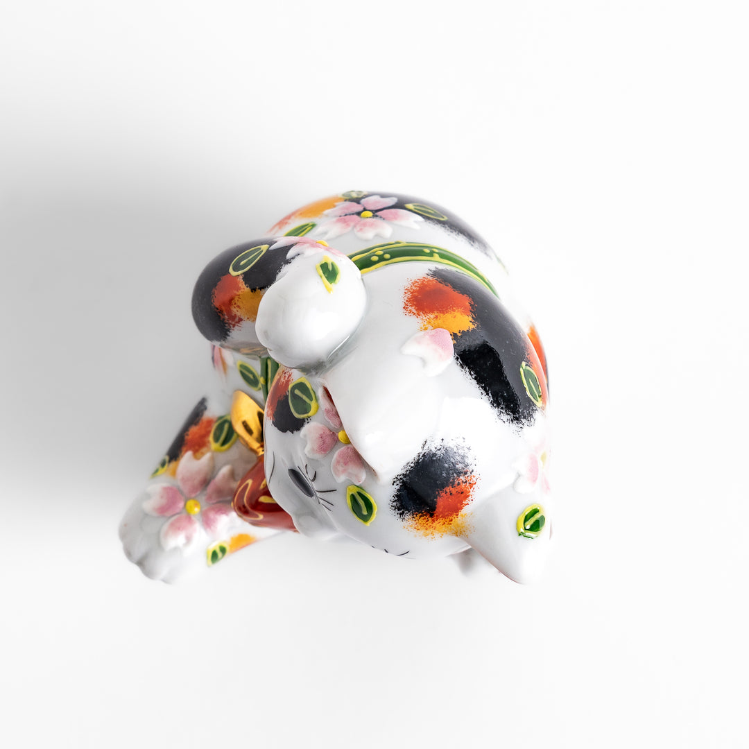 Colorful Calico Maneki Neko figurine holding a lucky sea bream, adorned with floral and green leaf patterns, symbolizing good fortune and prosperity in Japanese culture.