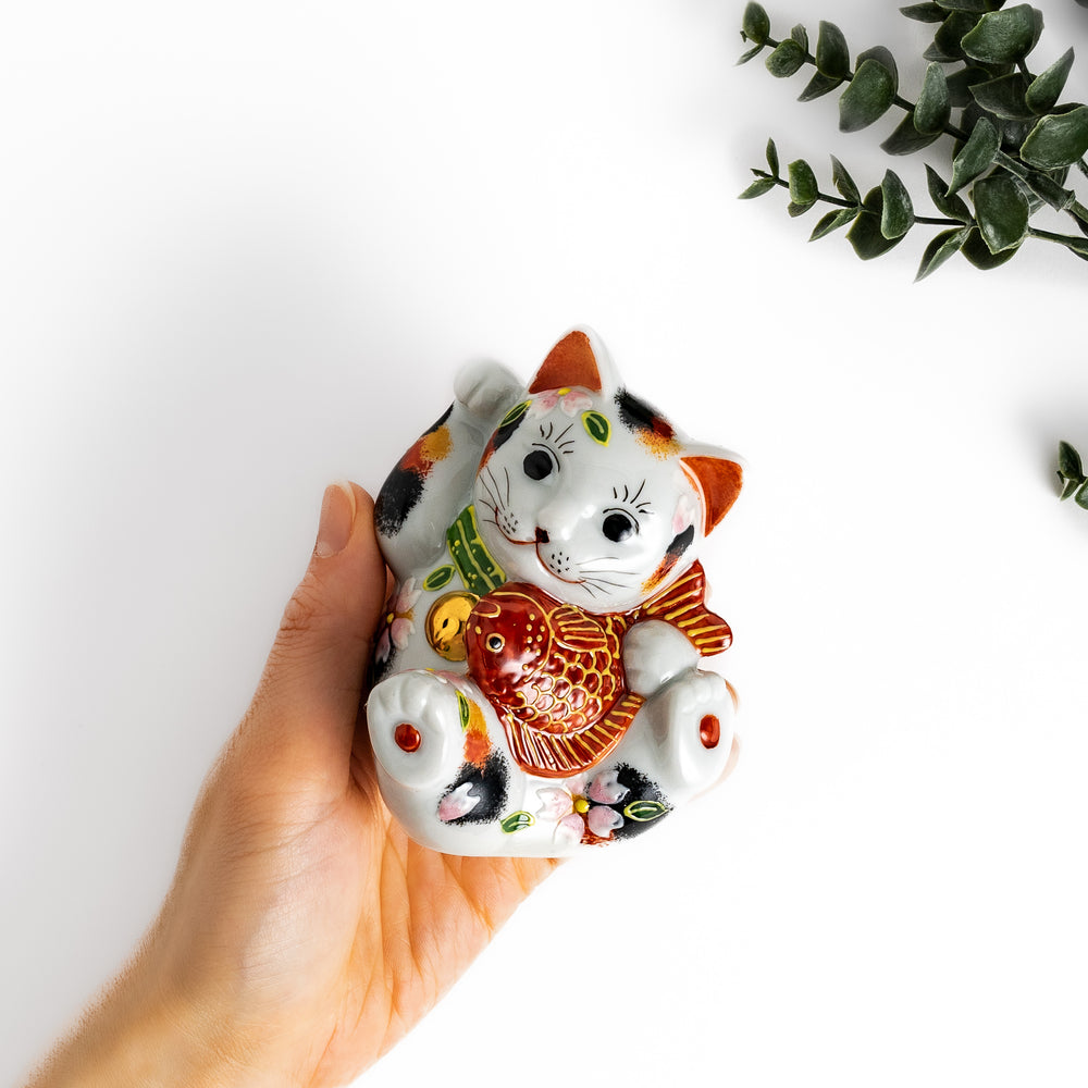 Colorful Calico Maneki Neko figurine holding a lucky sea bream, adorned with floral and green leaf patterns, symbolizing good fortune and prosperity in Japanese culture.