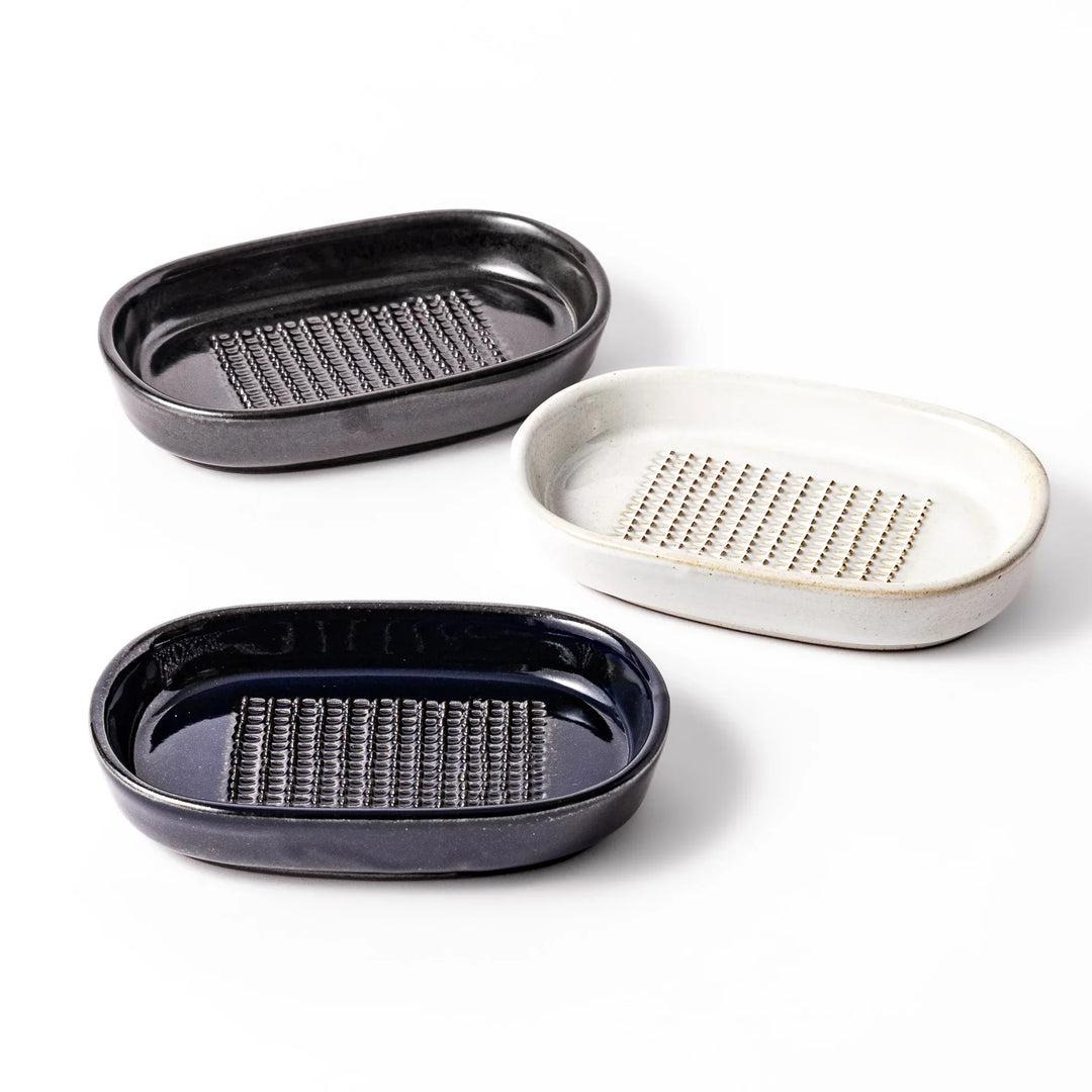 Three ceramic ginger grater dishes in black, white, and deep blue, each featuring raised teeth and a shallow design for efficient grating.