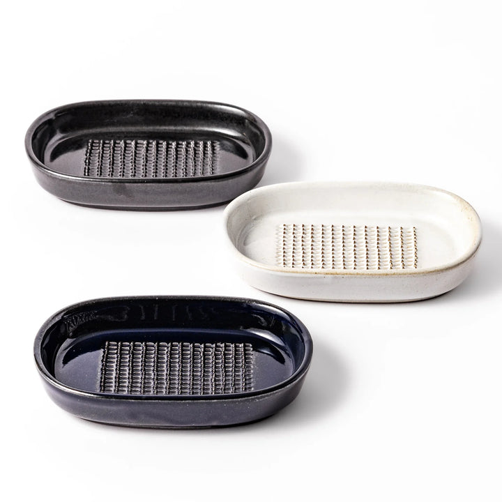 Three ceramic ginger grater dishes in black, white, and deep blue, each featuring raised teeth and a shallow design for efficient grating.
