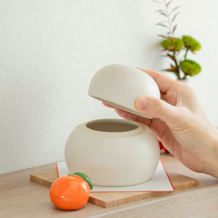 A person lifting the top of a white ceramic Kagami Mochi jar revealing a functional storage compartment inside