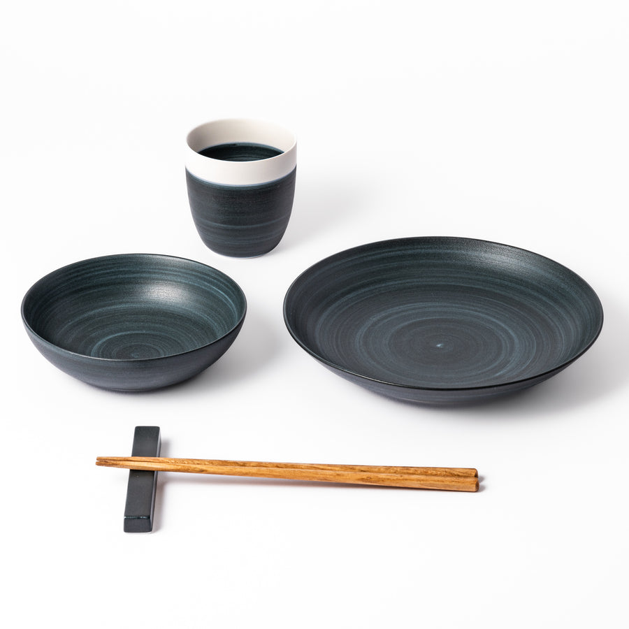 Japandi-style table setting in deep navy tones featuring a matching salad plate, bowl, tea cup, and chopsticks with rest.