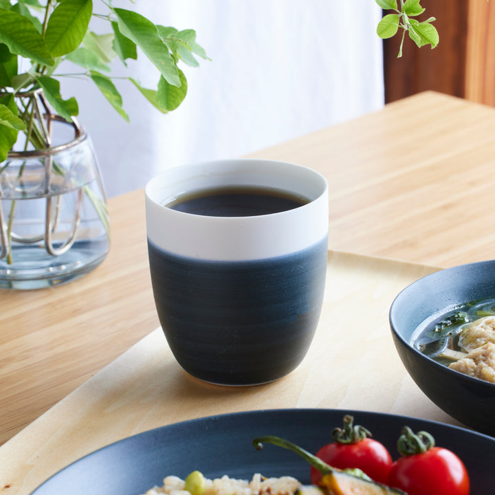 Cozy Japandi Tea Cup in a two-tone design, featuring dark navy and white, placed on a wooden table beside a meal and fresh greens.