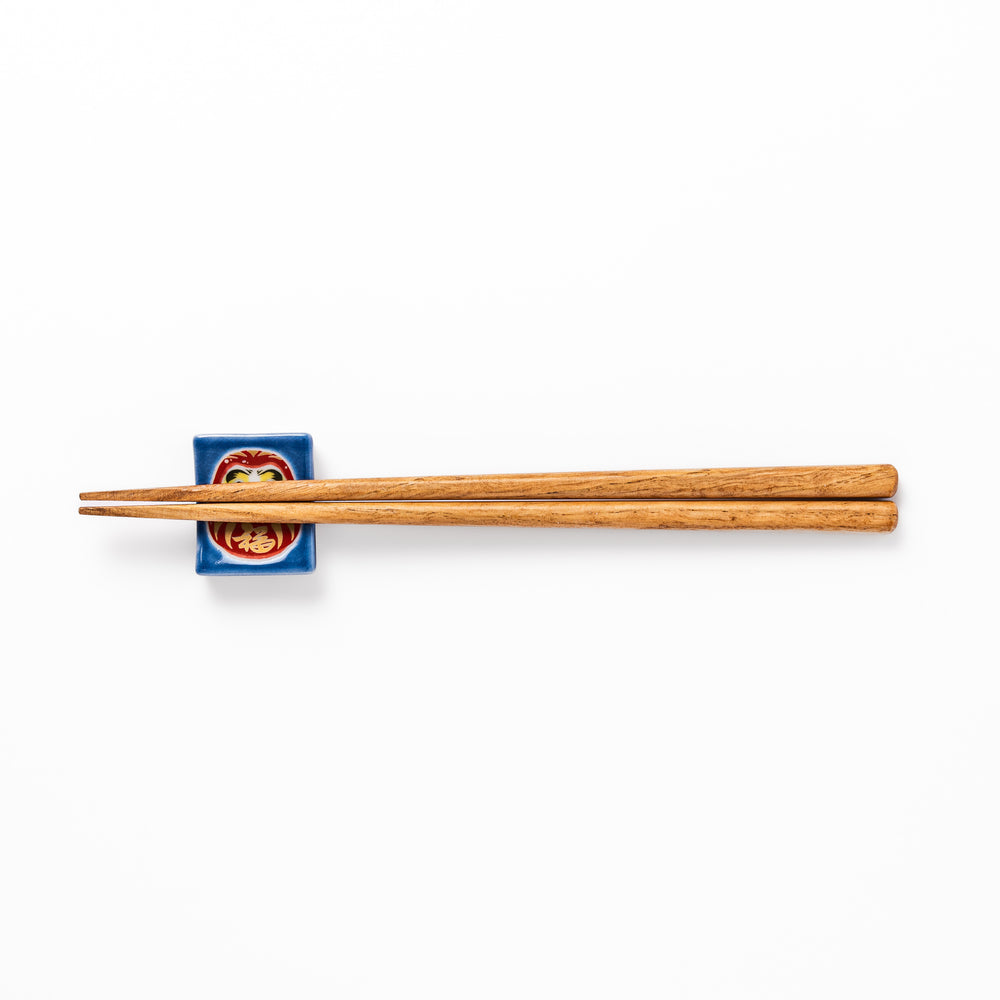 Rectangular Japanese ceramic chopstick rest featuring a red Daruma doll on a blue background, symbolizing perseverance, luck, and good fortune.