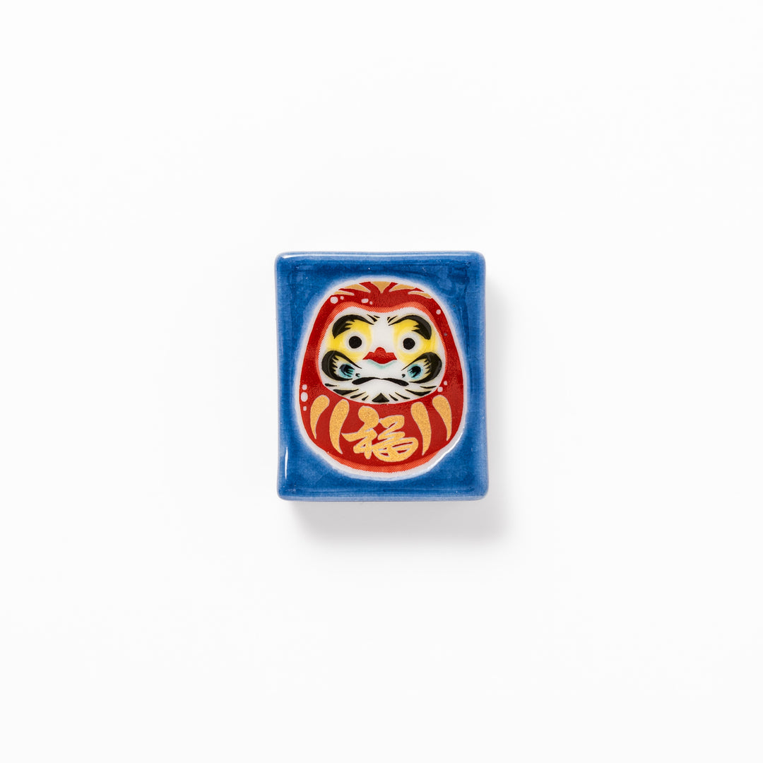 Rectangular Japanese ceramic chopstick rest featuring a red Daruma doll on a blue background, symbolizing perseverance, luck, and good fortune.