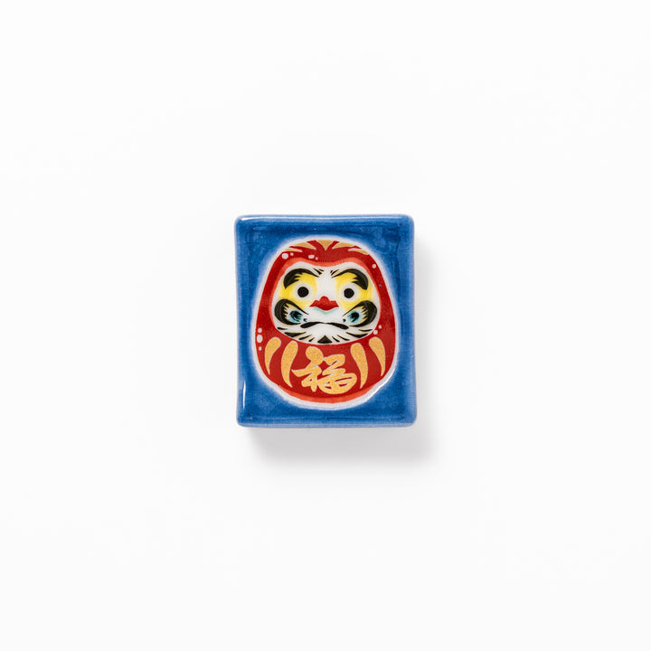 Rectangular Japanese ceramic chopstick rest featuring a red Daruma doll on a blue background, symbolizing perseverance, luck, and good fortune.