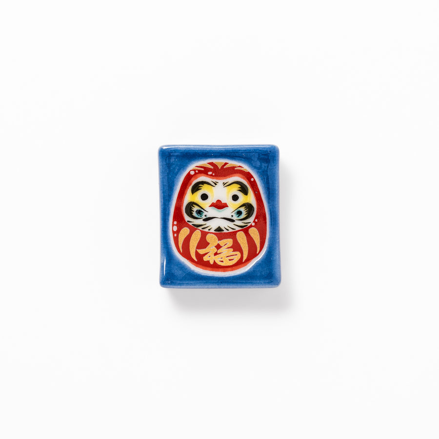 Rectangular Japanese ceramic chopstick rest featuring a red Daruma doll on a blue background, symbolizing perseverance, luck, and good fortune.