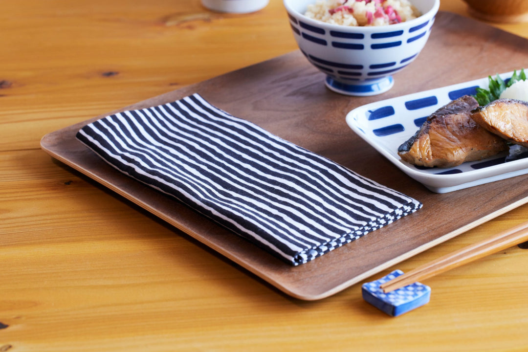 Striped black and white cloth on a wooden tray beside blue and white Japanese tableware with grilled fish and rice