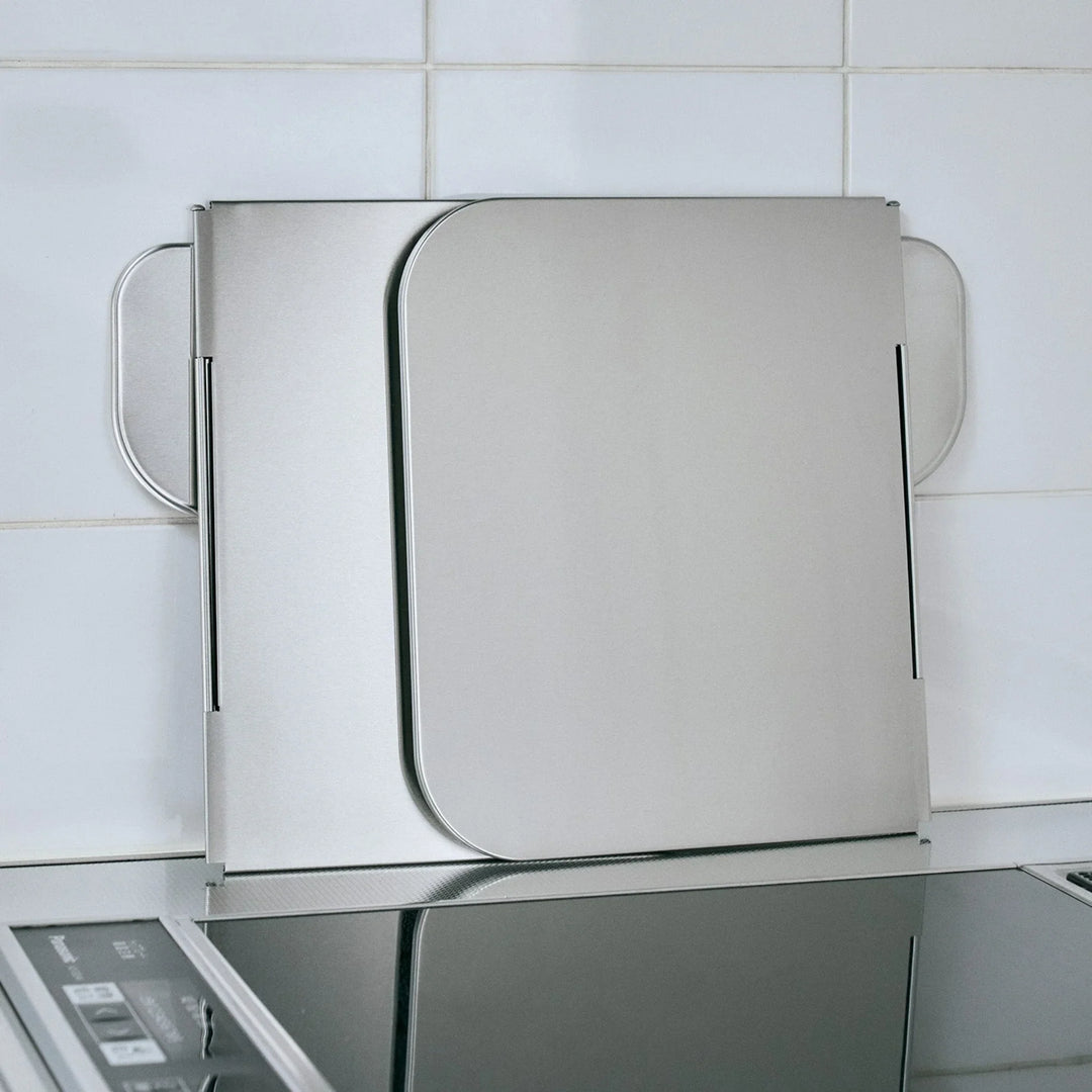 Foldable stainless steel stove splatter guard folded compactly and leaning against a tiled kitchen wall