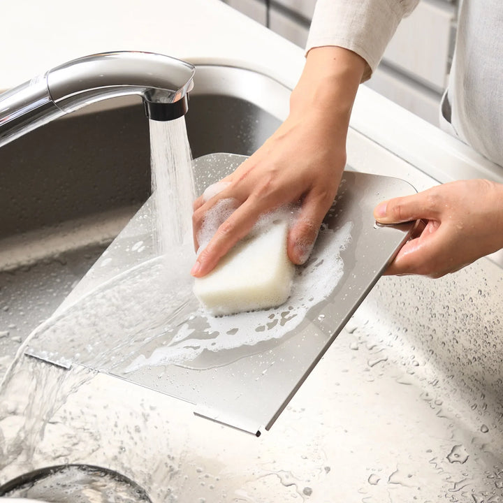 Person washing a stainless steel splatter guard panel with a soapy sponge in a kitchen sink