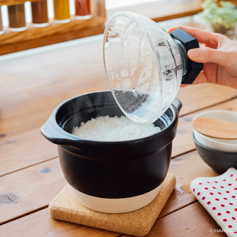 Lifting clear glass lid of hario rice cooker donabe to reveal steaming freshly cooked white rice japanese clay pot