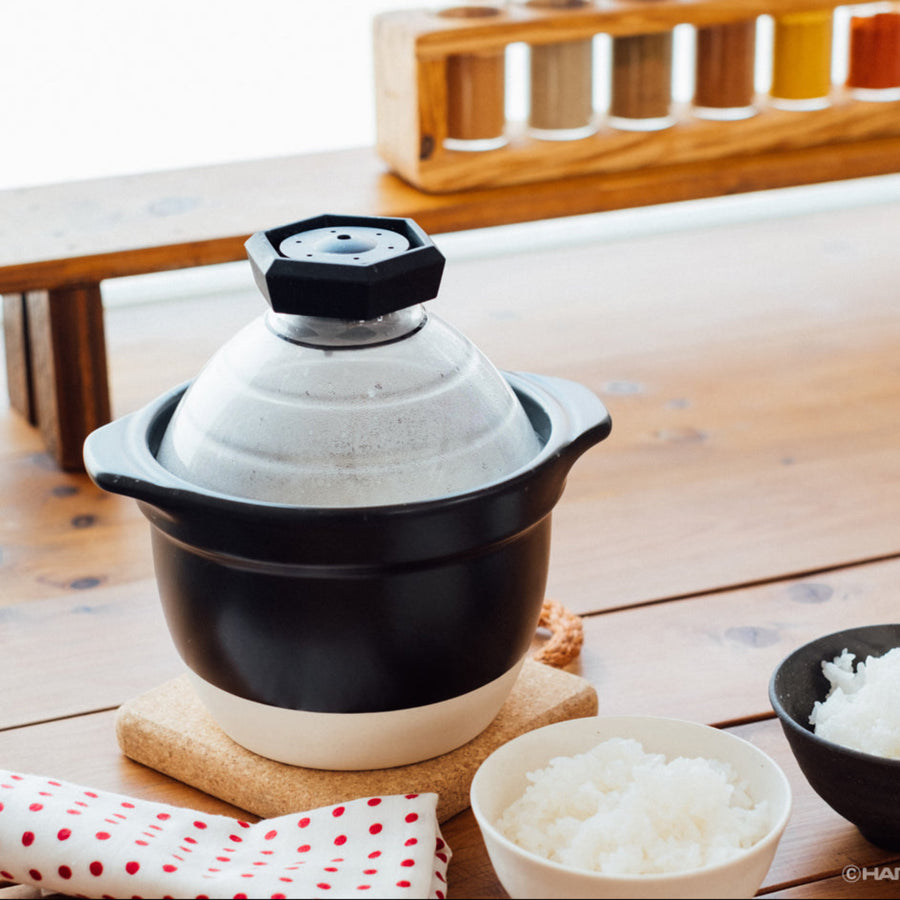 Hario glass lid rice cooker donabe clay pot sitting on wooden table with bowls of freshly cooked white rice