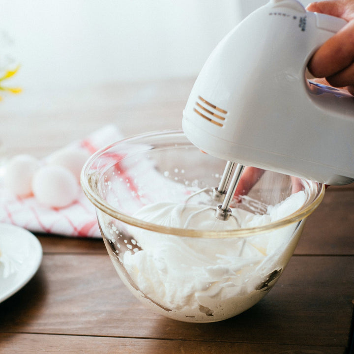 Whipping white cream with electric hand mixer in hario heatproof glass bowl on wooden table baking preparation