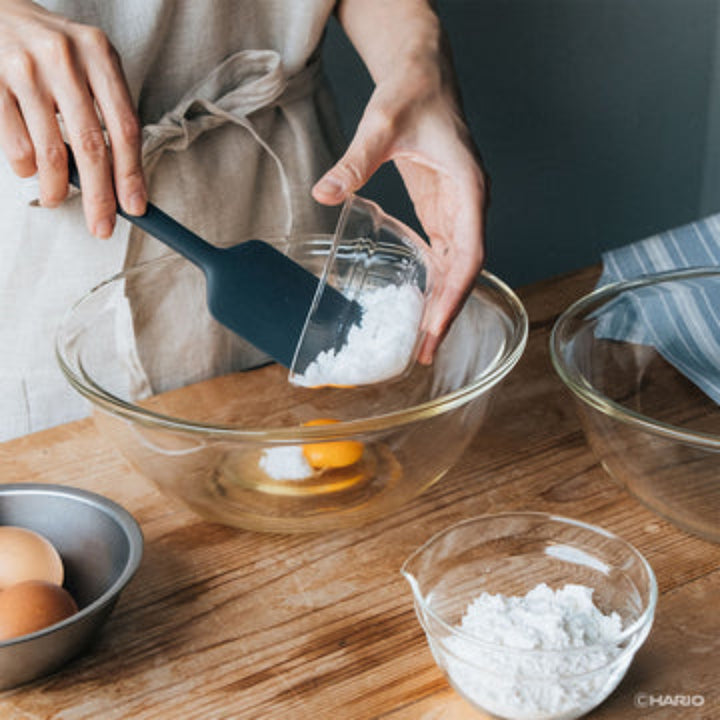 Adding sugar to egg yolk inside hario heatproof glass mixing bowl with spatula during sweet dessert baking prep